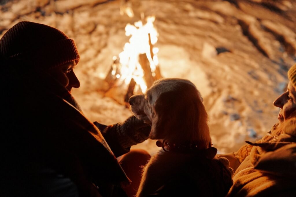 A couple enjoys a warm winter night by a bonfire, accompanied by their dog amidst a snowy setting.
